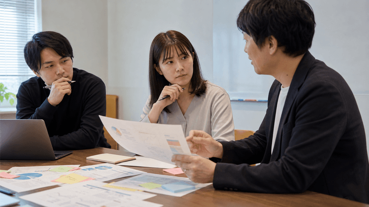 地方企業の研修担当者と現場リーダーが、研修資料と付箋を見ながら生成AI研修後の質問対応フローを話し合っている様子