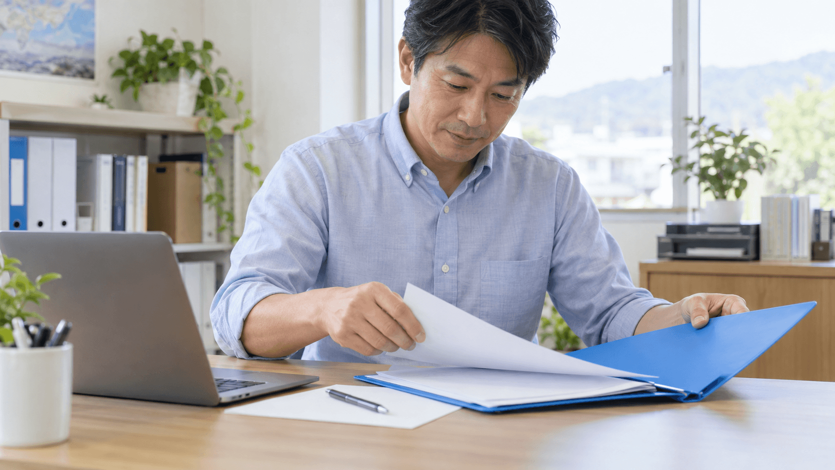 明るいオフィスで、担当者がノートPCの横に青いファイルと紙を広げ、手作業で業務を進める準備をしている様子