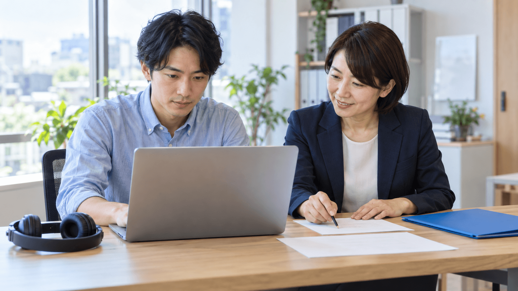 明るい事務所で社員がノートPCを操作し、隣の講師が演習用紙を確認している。机には録画学習用のヘッドホンと青いフォルダーが置かれている