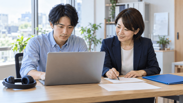 明るい事務所で社員がノートPCを操作し、隣の講師が演習用紙を確認している。机には録画学習用のヘッドホンと青いフォルダーが置かれている