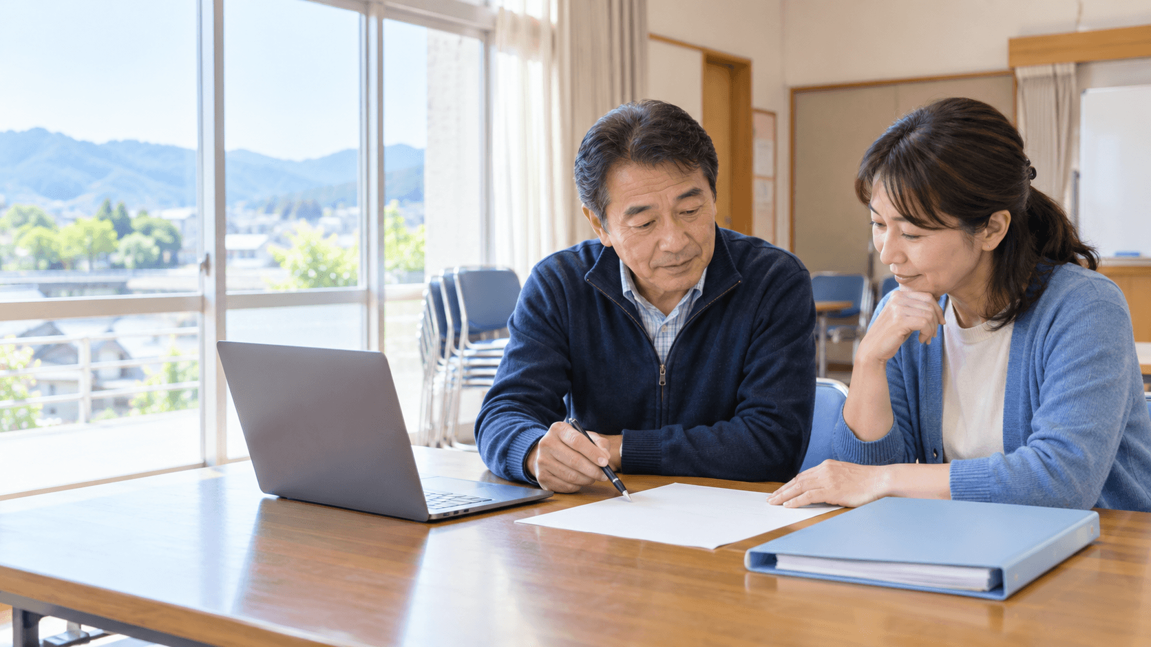 明るい地域の集会所で、二人の地域団体担当者がノートPC、無地の名簿用紙、青いファイルを机に置き、情報の保管方法を相談している様子