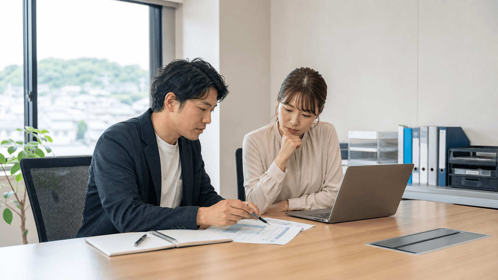 地方企業の会議室で2人の担当者がノートPCと配布資料を見ながら、AI議事録ツール導入前の会議ルール整理を相談している様子