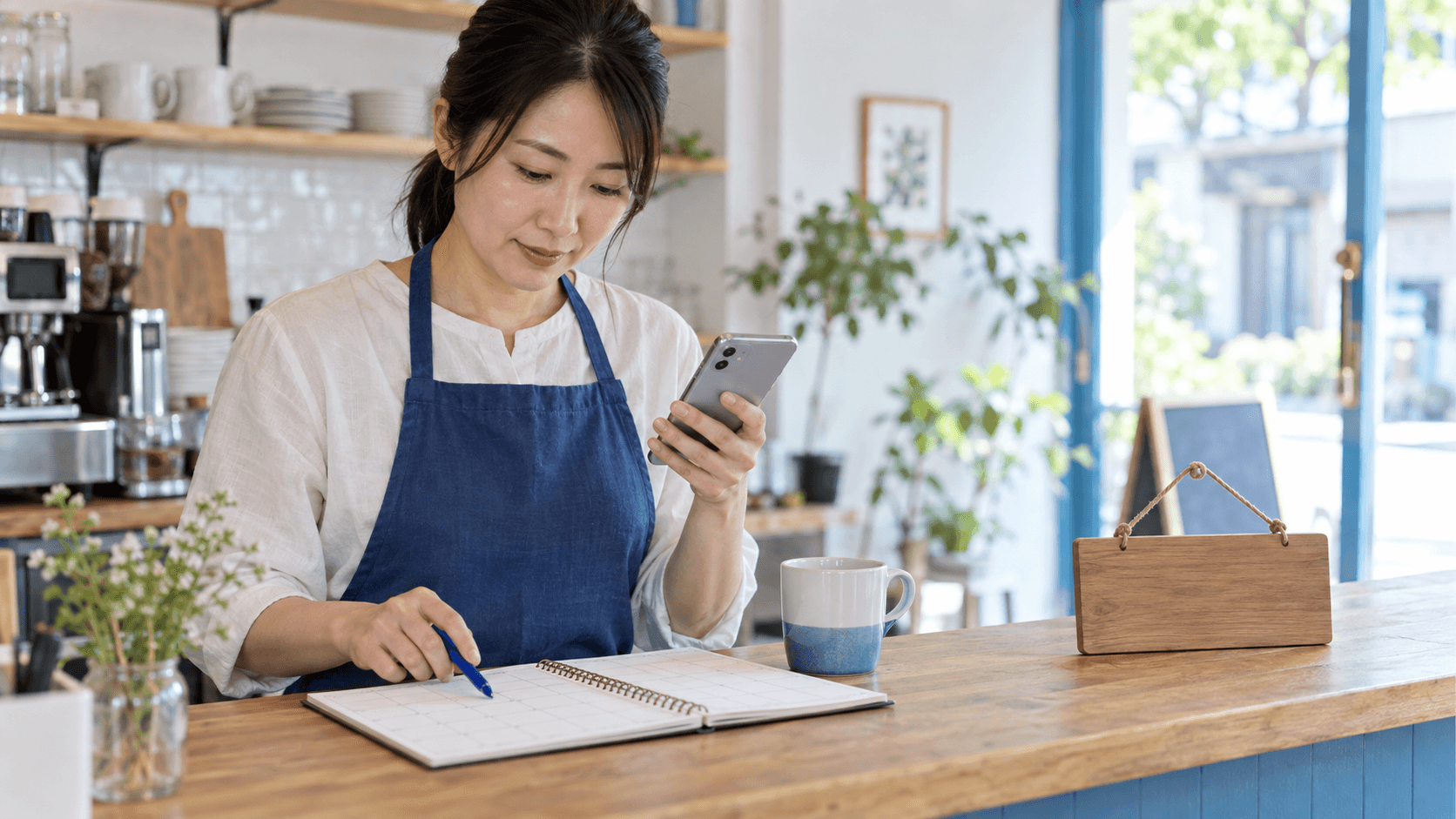 明るいカフェのカウンターで、青いエプロンの店主が紙の予定表とスマートフォンを見比べて営業予定を確認する様子