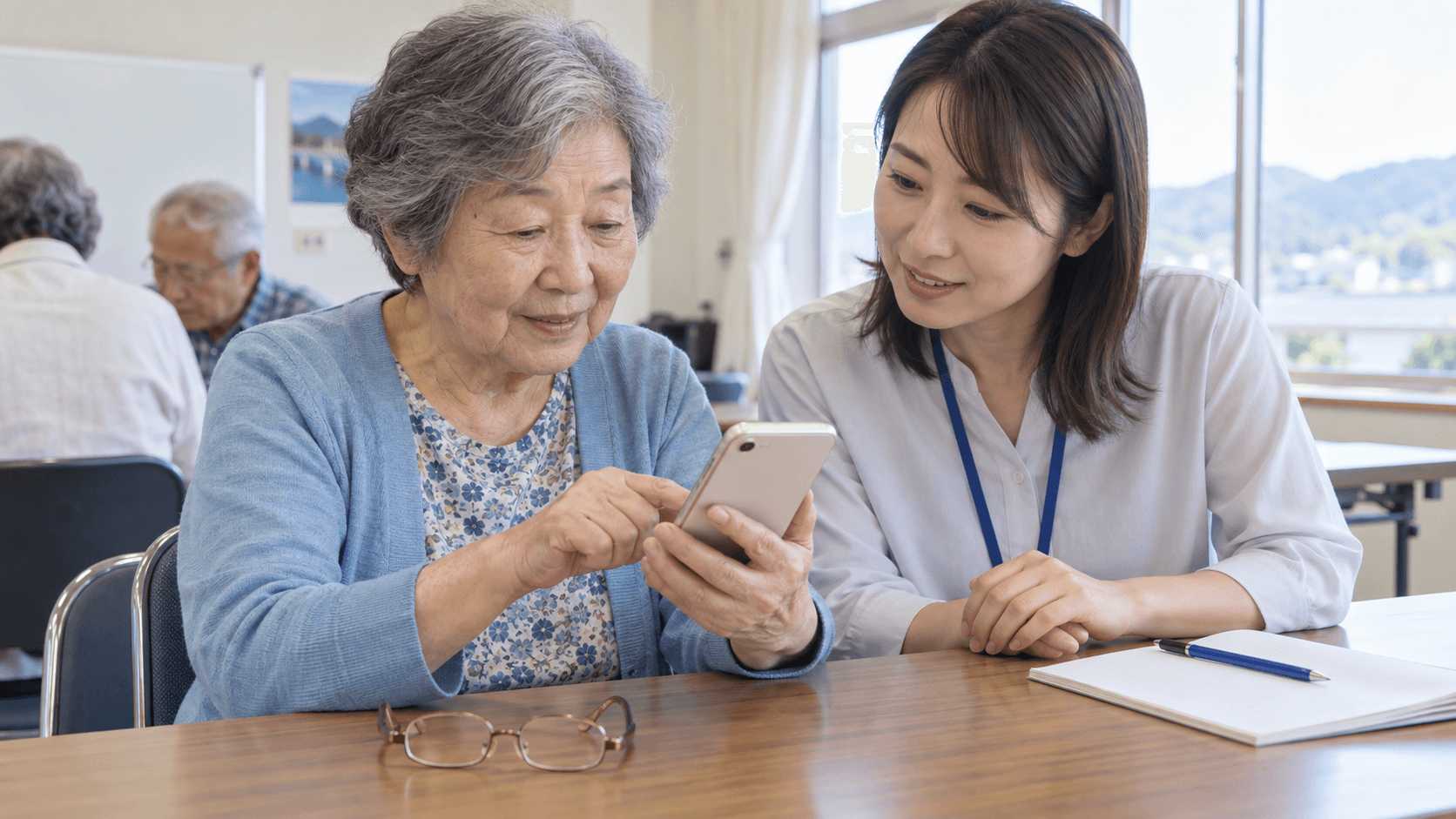 地域の明るい教室で、高齢の女性が自分のスマホを持って操作し、隣の講師が画面の見やすさを一緒に確認している様子